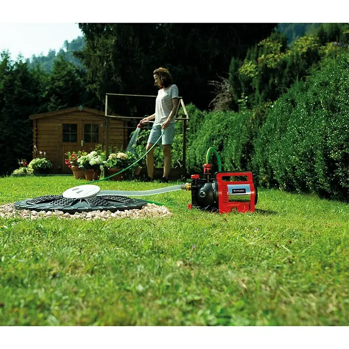 Einhell Gartenpumpe GC-GP 1250 N - Leistung: 1.200 W, Max. Fördermenge: 5.000 l/h - Ein roter und schwarzer Einhell Hauswasserautomat steht auf einem Rasen, während eine Frau im Hintergrund mit einem Gartenschlauch gießt.