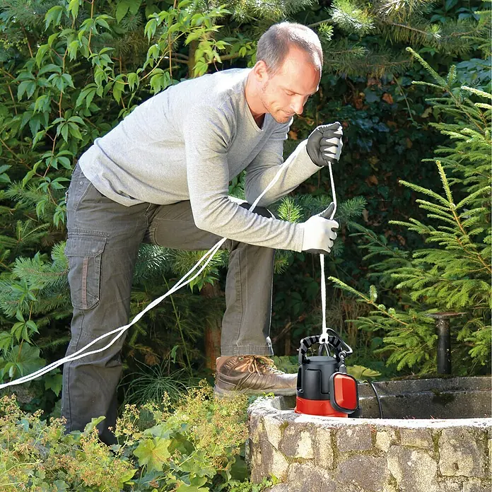 Einhell Schmutzwasserpumpe GC-DP 7835 - 780 W, Max. Fördermenge: 15.700 l/h, Eintauchtiefe: 7 m - Ein Mann lässt eine rote und schwarze Tauchpumpe mit einem Seil in einen Brunnen hinab. Er trägt Arbeitskleidung und Handschuhe.