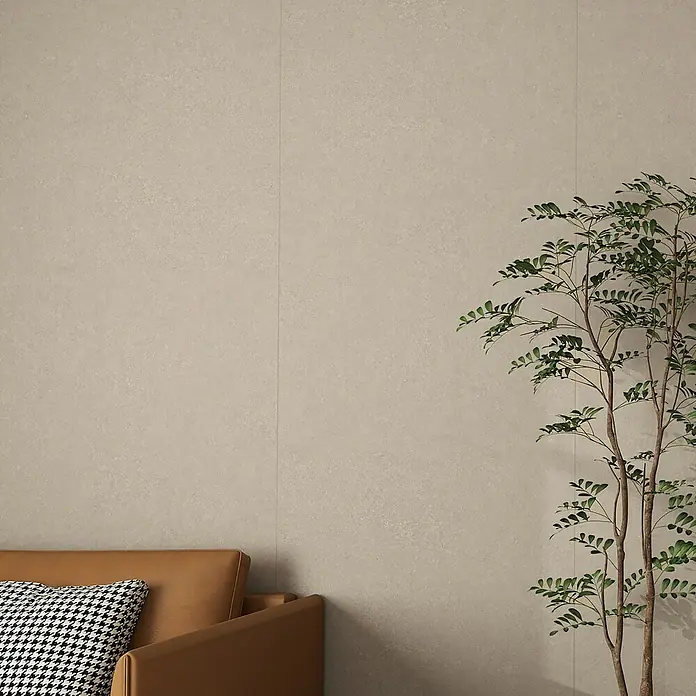 Wandpaneel Fliese Kampagne - 260 cm x 90 cm x 4 mm, Beige - Nahaufnahme eines braunen Sofas mit gemusterten Kissen vor einer beigen Wand, ergänzt durch eine elegante Zimmerpflanze.