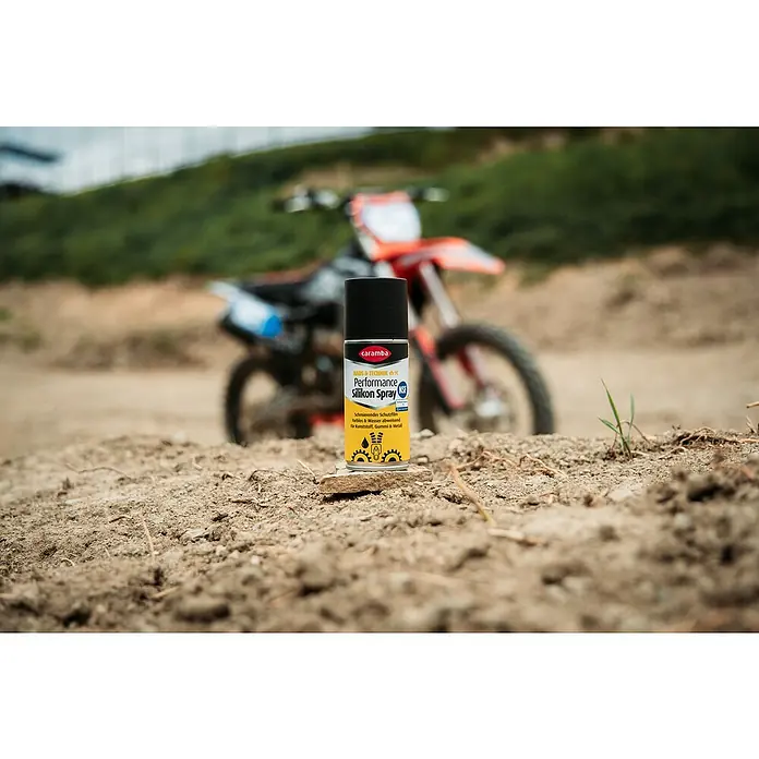 Caramba Silikonspray Performance Duo - 100 ml - Eine Dose Caramba Performance Silikon Spray vor einem unscharfen Motorrad auf erdigem Untergrund.