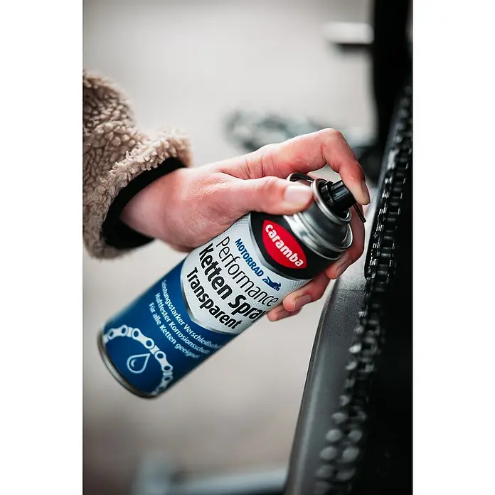 Caramba Kettenspray Performance - 300 ml - Kettenspray: Eine Hand sprüht Caramba Ketten Performance Spray Transparent auf eine Motorradkette, um sie zu schmieren und zu schützen.