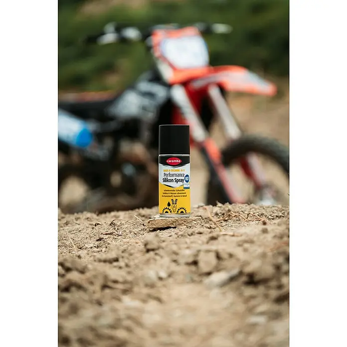 Caramba Silikonspray Performance Duo - 100 ml - Das Bild zeigt eine Dose Caramba Performance Silikon Spray vor einem unscharfen Motocross-Motorrad im Hintergrund.
