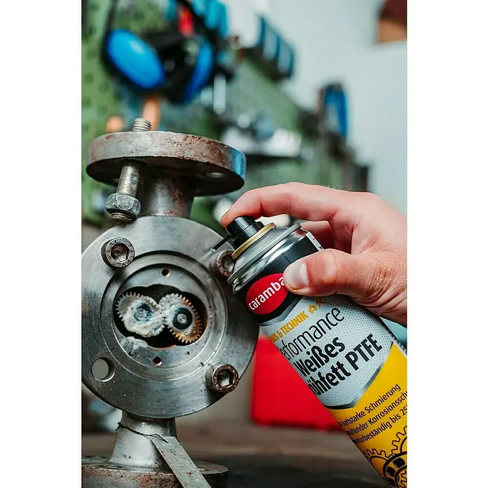 Caramba Mehrzweckfett Performance - 250 ml - Schmierende Hand: Eine Dose Caramba Performance Weißes Sprühfett PTFE wird auf ein Maschinenteil mit Zahnrädern aufgetragen, um die Schmierung zu verbessern.