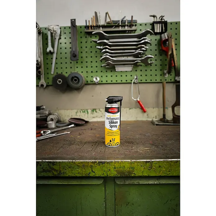 Caramba Silikonspray Performance Duo - 300 ml - Eine Dose Caramba Performance Silikonspray steht auf einer Werkbank vor einer grünen Werkzeugwand, ideal für Wartung und Pflege.