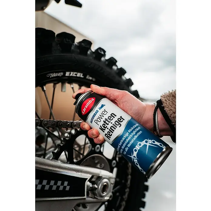Caramba Kettenreiniger - 300 ml - Eine Dose Caramba Motorrad Kettenreiniger wird vor dem Hintergrund eines Motorradreifens gehalten.