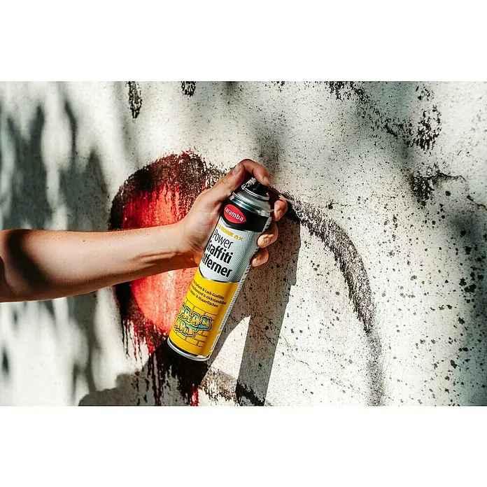 Caramba Graffiti-Entferner - 400 ml - Eine Hand hält eine Dose Caramba Power Graffiti Entferner vor einer Wand mit Graffiti, um diese zu entfernen.