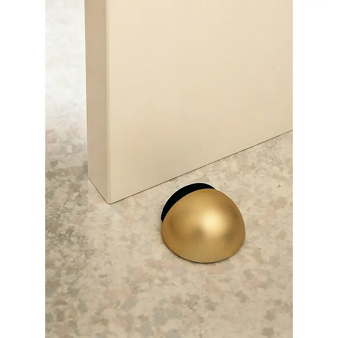 Inofix Tope de puerta 2029 - Plástico, Marrón - Primer plano de un elegante tope de puerta dorado junto a una pared color crema sobre un suelo moteado.