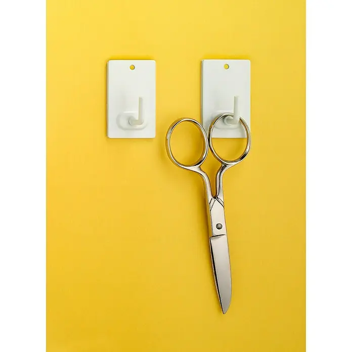 Inofix Colgador adhesivo 2040 - Acero inoxidable, Blanco - Tijeras plateadas afiladas que cuelgan de dos ganchos adhesivos blancos fijados a una pared amarilla, listas para usar de inmediato.