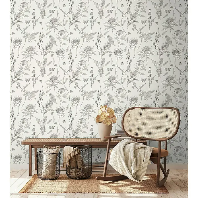 A.S. Création Papel pintado de vellón Flores y Animales - Beige, Floral, 10,05 x 0,53 m - Elegante interior con una mecedora de ratán, cestas y un banco decorativo frente a un papel pintado floral.