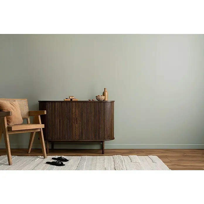 swingcolor Wandfarbe Comfort Home - Stone Grey, Stumpfmatt, 2,5 l, Konservierungsmittelfrei - Elegantes Interieur mit einem dunklen Holzschrank, einem hellen Holzstuhl mit Kissen und einem gemusterten Teppich, der eine warme Atmosphäre schafft.
