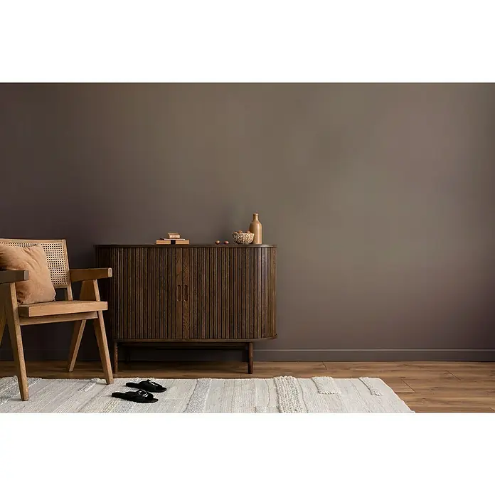 swingcolor Wandfarbe Comfort Home - Magic Wall, Stumpfmatt, 2,5 l, Konservierungsmittelfrei - Elegantes Sideboard aus dunklem Holz mit geriffelten Türen, dekoriert mit einer Flasche und einer Schale. Im Vordergrund ein Holzstuhl mit Kissen.