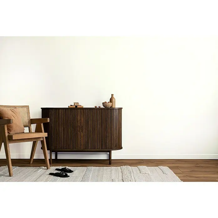 swingcolor Wandfarbe Comfort Home Mineral-Innenfarbe - Snow White, Stumpfmatt, 5 l, Konservierungsmittelfrei - Einrichtung mit einem dunklen Holzschrank mit geriffelten Türen, dekoriert mit einer Flasche und einer Schale, neben einem Stuhl auf einem gemusterten Teppich.