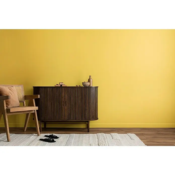 swingcolor Wandfarbe Comfort Home - Golden Dust, Stumpfmatt, 2,5 l, Konservierungsmittelfrei - Helles Interieur mit gelber Wand, einem Holzschrank, einem Stuhl mit Kissen und einem hellen Teppich mit dunklen Slippern.