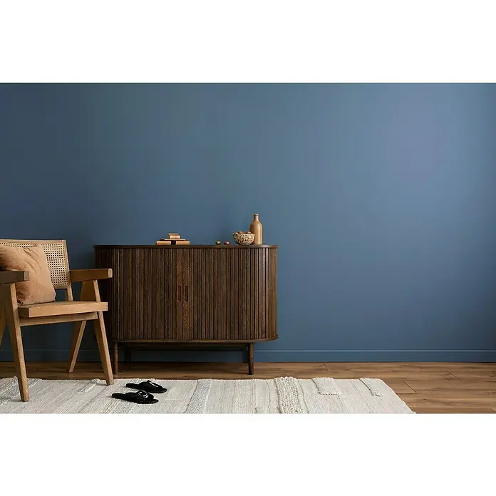 swingcolor Wandfarbe Comfort Home - Blue Sea, Stumpfmatt, 2,5 l, Konservierungsmittelfrei - Elegantes Interieur mit einem dunklen Holzschrank, einem hellen Holzstuhl und einem stilvollen Teppich vor einer blauen Wand.