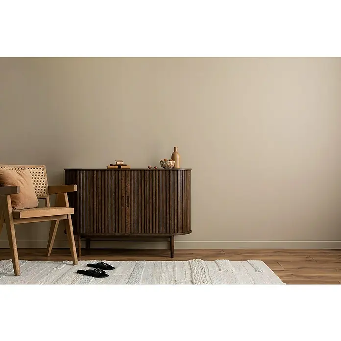 swingcolor Wandfarbe Comfort Home - Beige Stone, Stumpfmatt, 1 l, Konservierungsmittelfrei - Ein stilvolles Interieur mit einem dunklen Holzschrank, einem Rattan-Stuhl und einem hellen Teppich, das eine gemütliche Atmosphäre schafft.