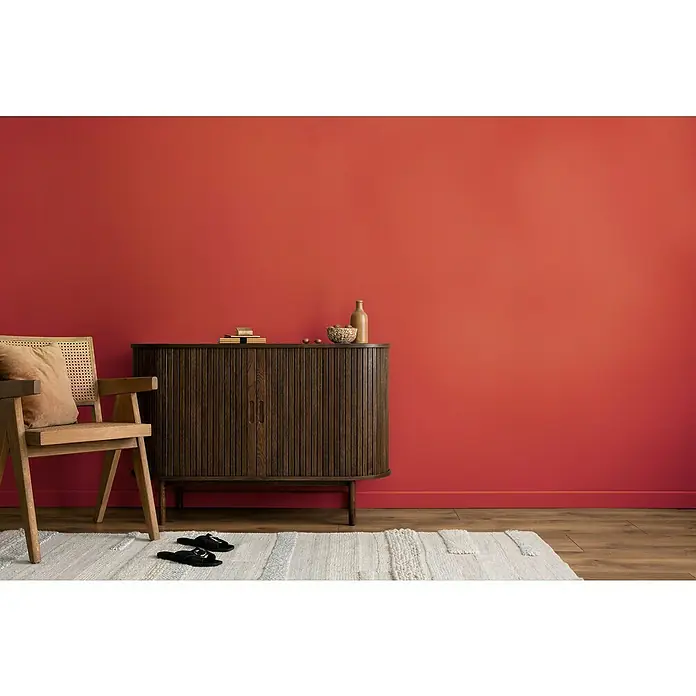 swingcolor Wandfarbe Comfort Home - Moon Red, Stumpfmatt, 1 l, Konservierungsmittelfrei - Das Bild zeigt ein Sideboard aus Holz mit einer Flasche und Dekorationen darauf, neben einem Stuhl auf einem hellen Teppich.