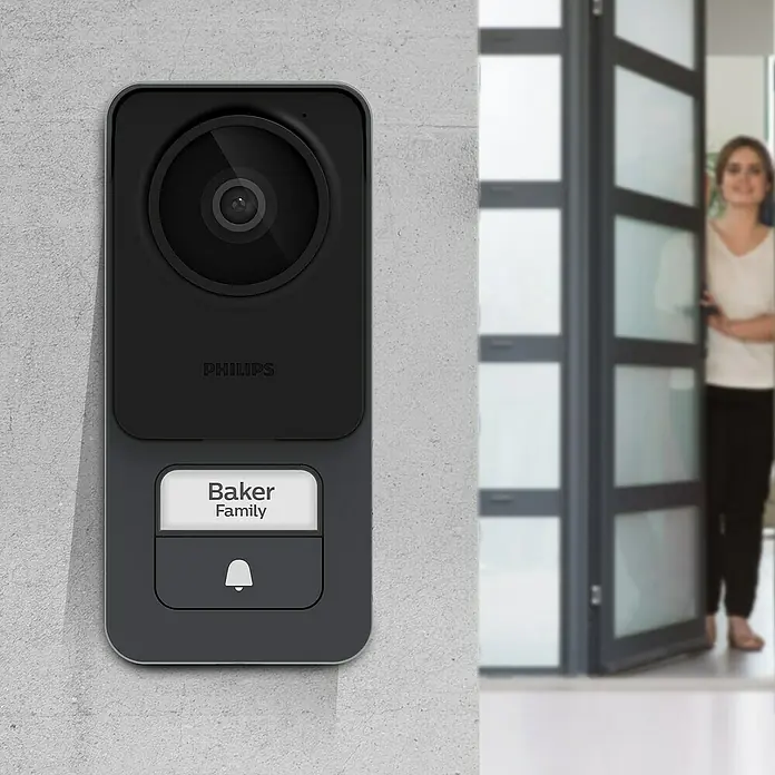 Philips WelcomeEye Farb-Video-Türsprechanlage Lite 2 - 1-Familienhaus - Nahaufnahme einer Philips Türklingel mit Kamera und Namensschild "Baker Family" an einer Wand, neben einer teilweise geöffneten Tür.