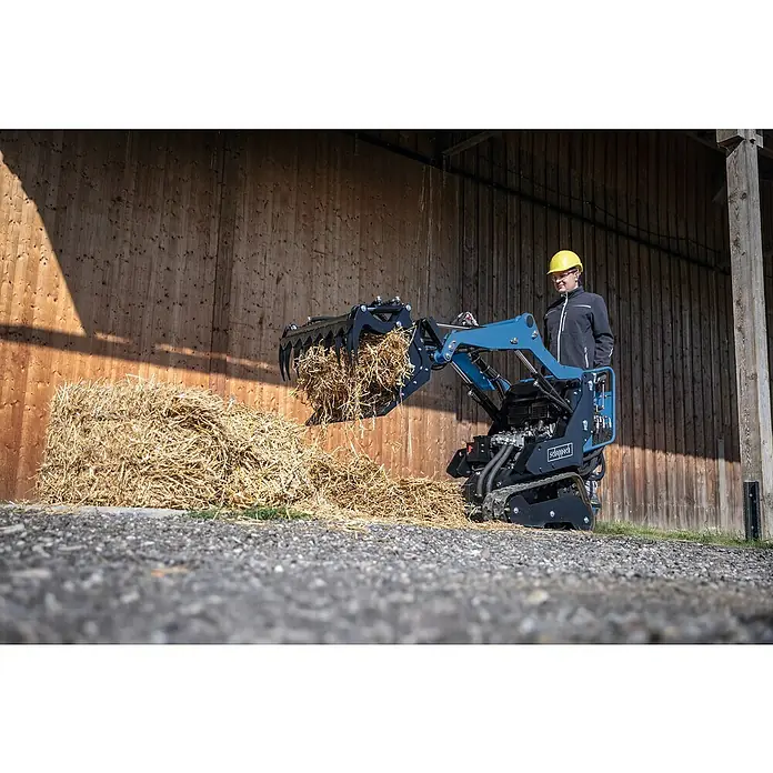 Scheppach Greifschaufel - 80 l, Passend für: Scheppach Kompaktlader MKL730 - Der Scheppach Hoflader mit Schaufel voller Stroh und einem Mann mit gelbem Helm vor einem Holzgebäude.