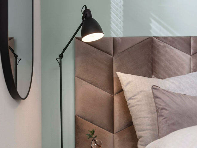 Graue Wandpolster hinter dem Bett, daneben eine Stehleuchte