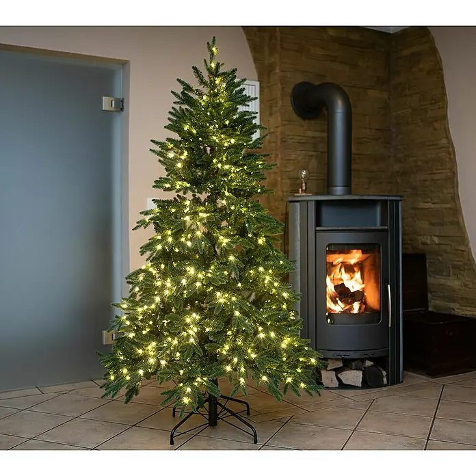 50 NRTH Künstlicher Weihnachtsbaum Sherwood - Höhe: 210 cm, Grün, LED-Beleuchtung - Geschmückter Weihnachtsbaum mit Lichtern neben einem Kamin. Der künstliche Weihnachtsbaum sorgt für festliche Stimmung im Raum.