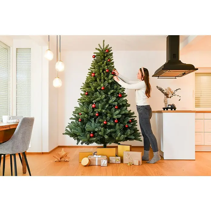 50 NRTH Künstlicher Weihnachtsbaum Vermont - Höhe: 240 cm, Grün, Standfuß - Frau schmückt einen großen, üppigen Weihnachtsbaum mit roten Kugeln, umgeben von goldenen und weißen Geschenken auf einem warmen Holzboden.