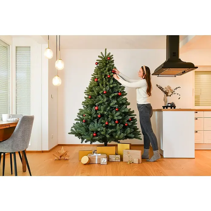 50 NRTH Künstlicher Weihnachtsbaum Vermont - Höhe: 210 cm, Grün, Standfuß - Eine Frau schmückt einen Weihnachtsbaum mit roten Kugeln, umgeben von Geschenken, in einem hellen, modern eingerichteten Raum. Festliche Dekoration.