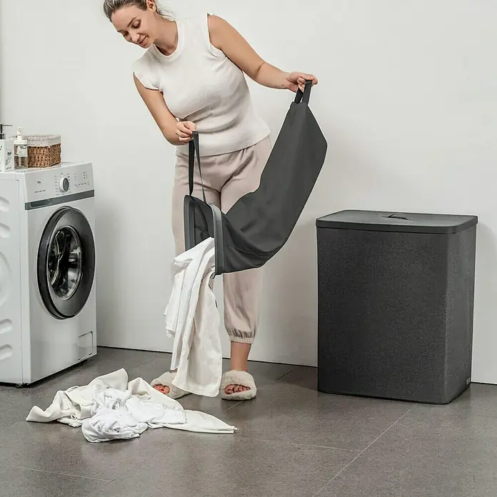 Store It Premium Wäschesammler Ava - 67 x 56 x 40 cm, Schwarz - Frau leert einen grauen Wäschesack neben einer Waschmaschine und einem Wäschekorb, wobei der Fokus auf dem Wäschesack liegt.