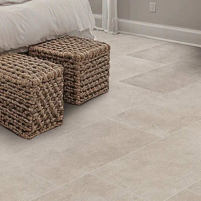 Feinsteinzeugfliese Rimini - 30,5 x 61 cm, Beige, Matt - Nahaufnahme von zwei braunen, gewebten Hockern auf hellgrauem Steinzeugboden neben einem Bett.