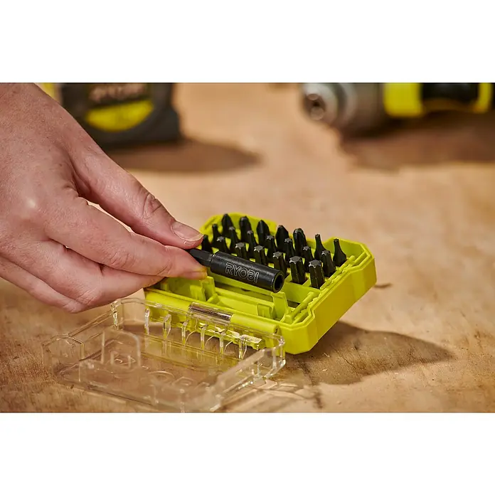 Ryobi Bit-Set RARXSD22 - Bitlänge: 25 mm, 22 -tlg. - Eine Hand hält einen Ryobi Bithalter über eine gelbe Bit-Box, die mit diversen Schrauberbits gefüllt ist. Werkzeug auf Holztisch.