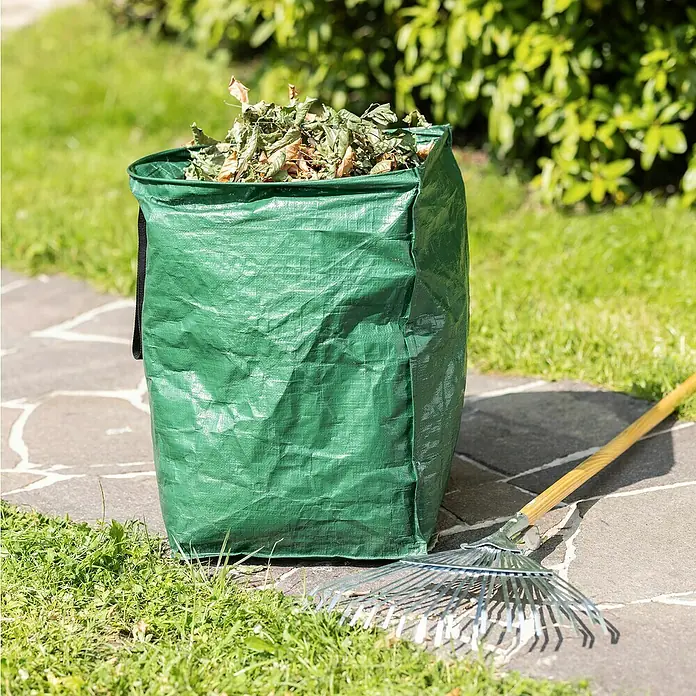 Windhager Gartenabfallsack - 150 l, Grün - Ein grüner Gartensack gefüllt mit Blättern steht neben einem Rechen auf einem gepflasterten Weg. Ideal für die Gartenarbeit!