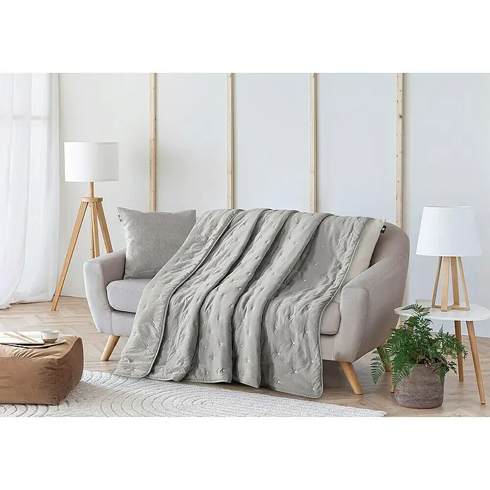 Edredón Bordado Estrellas - 180 x 260 cm, Poliéster, Gris - Una manta gris acolchada con un patrón de estrellas está colocada sobre un sofá beige. La manta añade un toque de elegancia y comodidad a la habitación.