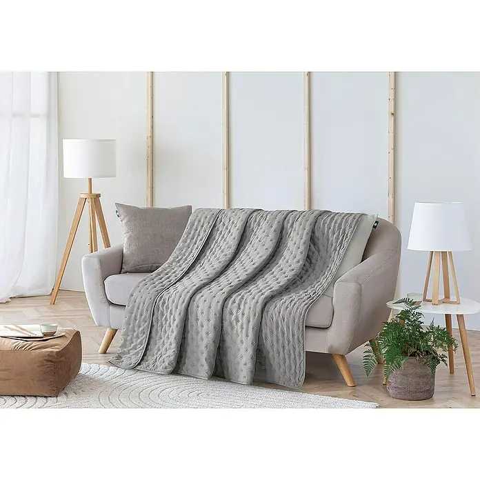 Edredón Corinto - 180 x 260 cm, Poliéster, Gris - Un tiro acolchado gris está elegantemente drapeado sobre un sofá beige, con una almohada a juego y lámparas elegantes en el fondo.
