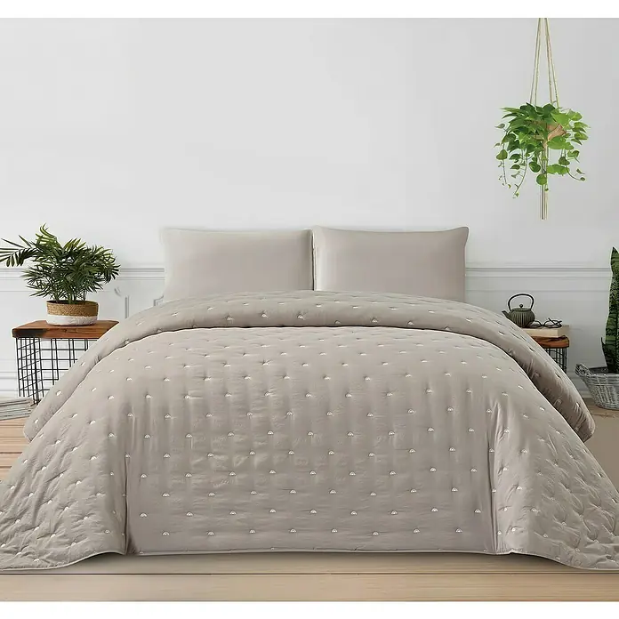 Edredón Bordado Sello - 220 x 250 cm, Poliéster, Taupe - Cubrecama beige con puntos bordados blancos y fundas de almohada a juego en una cama con dos mesitas de noche y plantas.