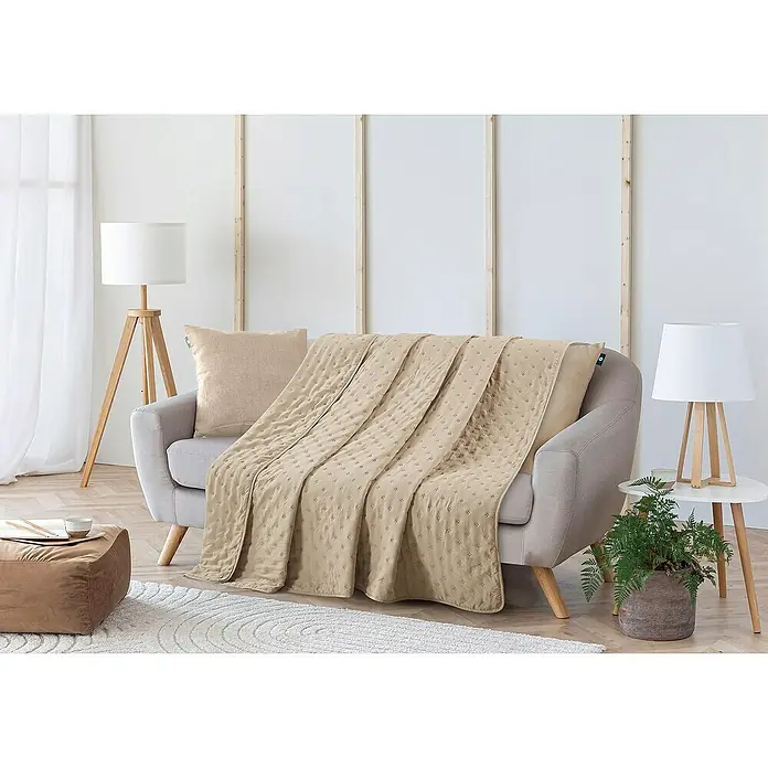Edredón Corinto - 180 x 260 cm, Poliéster, Taupe - Escena elegante con una manta acolchada beige sobre un sofá, complementada con una lámpara de pie y una mesa auxiliar con planta para un ambiente acogedor.