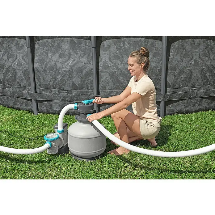 Bestway Frame-Pool Set APX365 - Ø x H: 671 x 132 cm, Dunkler Marmor, 40.377 l - Eine Frau bedient eine graue Poolsandfilteranlage mit weißen Schläuchen auf grünem Rasen vor einem grauen Pool.
