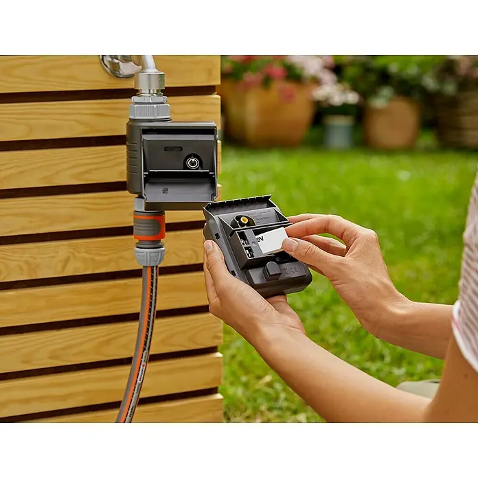 Gardena Bewässerungscomputer Master - Bewässerungsdauer: 1 s - 99 min - Das Bild zeigt eine Person, die eine 9V-Batterie in ein Gardena Bewässerungssteuerungsgerät einlegt, das an einem Wasserhahn im Garten befestigt ist.