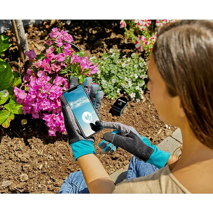 Gardena Smart system Bodenfeuchtesensor - Passend für: Gardena Smart System Sets - Eine Frau benutzt die Gardena Smart System App auf ihrem Smartphone im Garten, um ihre Pflanzen zu pflegen.