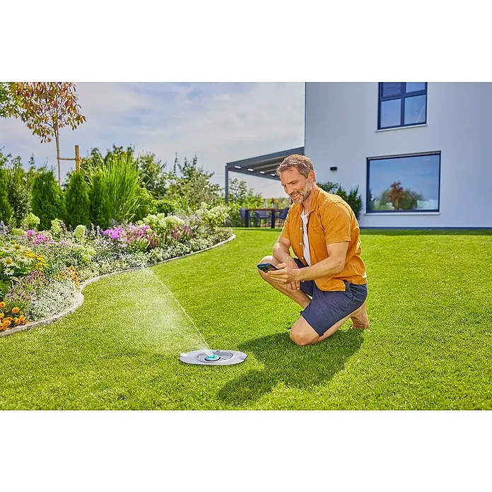 Gardena Sprinklersystem Versenkregner-Set AquaPrecise Basis - Max. Regnerfläche: 320 m², Reichweite ca.: 2 m - 10 m - Mann bedient smarten Rasensprenger mit dem Handy in einem gepflegten Garten. Das Gerät sorgt für eine gleichmäßige Bewässerung der Rasenfläche.