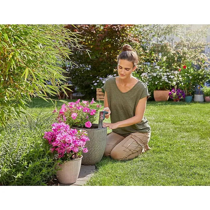 Gardena Smart system Bodenfeuchtesensor - Passend für: Gardena Smart System Sets - Eine Frau kniet im Garten und gießt Blumen in einem Topf mit einem GARDENA-Produkt, umgeben von üppigem Grün.
