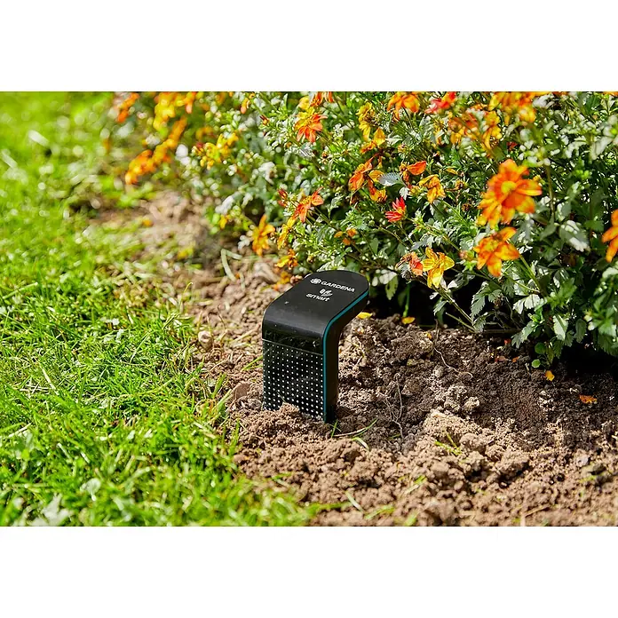 Gardena Smart system Bodenfeuchtesensor - Passend für: Gardena Smart System Sets - Das Bild zeigt einen Gardena Smart Sensor im Garten, der die Bodenfeuchtigkeit und Temperatur misst. Im Hintergrund blühende Pflanzen.
