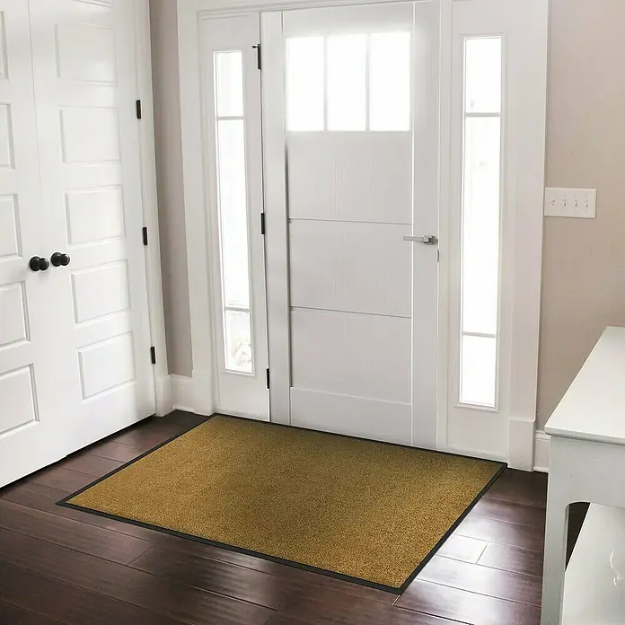 Fußmatte Rhine - Beige, 600 x 90 cm, 100 % Polyamid - Ein rechteckiger, goldfarbener Fußabtreter mit schwarzem Rand liegt vor einer weißen Tür auf einem dunklen Holzboden.
