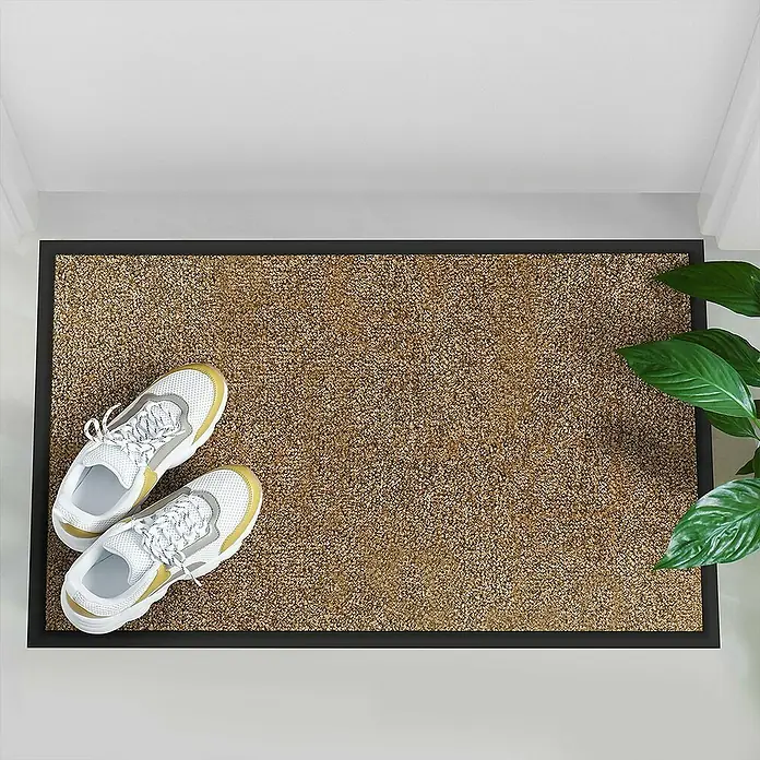 Fußmatte Rhine - Beige, 600 x 90 cm, 100 % Polyamid - Ein brauner Fußabtreter mit schwarzem Rahmen und weißen Schuhen darauf, neben einer grünen Pflanze.