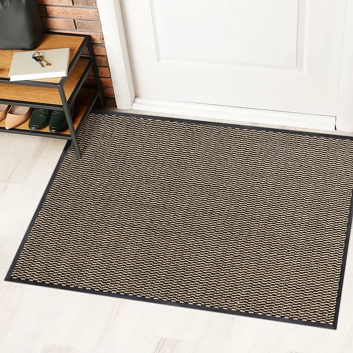 Fußmatte Sky Novo - Ocker/Schwarz, 120 x 90 cm, 100 % Polypropylen - Eine Fußmatte in Beige und Schwarz mit gewebtem Muster und dunklem Rand liegt vor einer weißen Tür. Ein Schuhregal steht daneben.