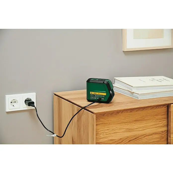 Bosch Kreuzlinienlaser Advanced Level 2G Set - Max. Arbeitsbereich: 15 m - Ein grüner Bosch-Laser-Entfernungsmesser wird auf einem Holzschrank aufgeladen. Der Stecker ist in eine Steckdose an der Wand gesteckt.