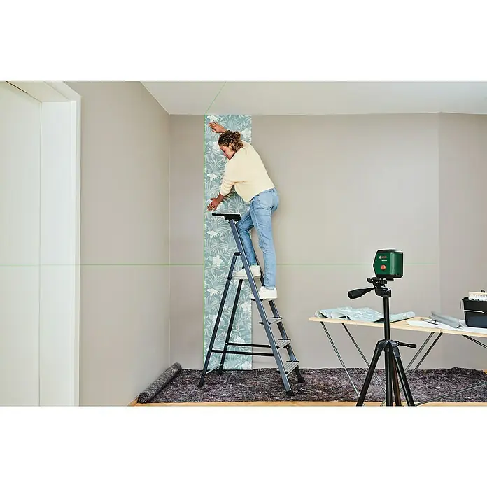 Bosch Kreuzlinienlaser Advanced Level 2G Set - Max. Arbeitsbereich: 15 m - Frau tapeziert Wand mit grünem Bosch Kreuzlinienlaser für präzise Ausrichtung, Leiter und Werkzeug bereit.