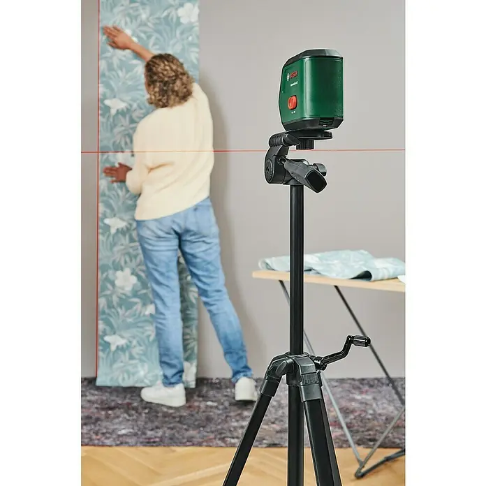 Bosch Universal Kreuzlinienlaser UniversalLevel 2 Set - Max. Arbeitsbereich: 12 m, Mit Stativ - Eine Frau tapeziert mit Hilfe des Bosch UniversalLevel 2, der auf einem Stativ steht und rote Laserlinien an die Wand projiziert.