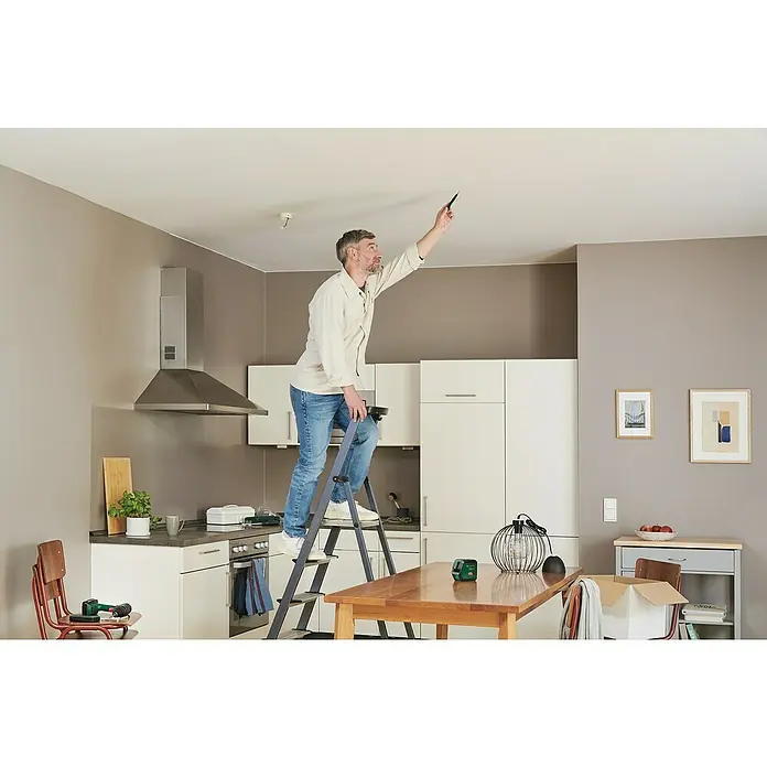 Bosch Kreuzlinienlaser Advanced Level 2G Set - Max. Arbeitsbereich: 15 m - Ein Mann steht auf einer Leiter und inspiziert die Decke mit einem Stift in der Hand. Helle Küche mit Esstisch im Hintergrund.