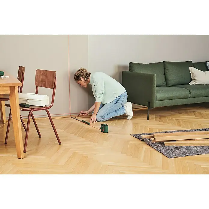 Bosch Kreuzlinienlaser UniversalLevel 2 - Max. Arbeitsbereich: 5 m - 20 m - Eine Frau kniet mit einem Bosch Laser-Nivelliergerät und markiert eine Linie an einer Wand, während sie ein kleines Lineal benutzt.