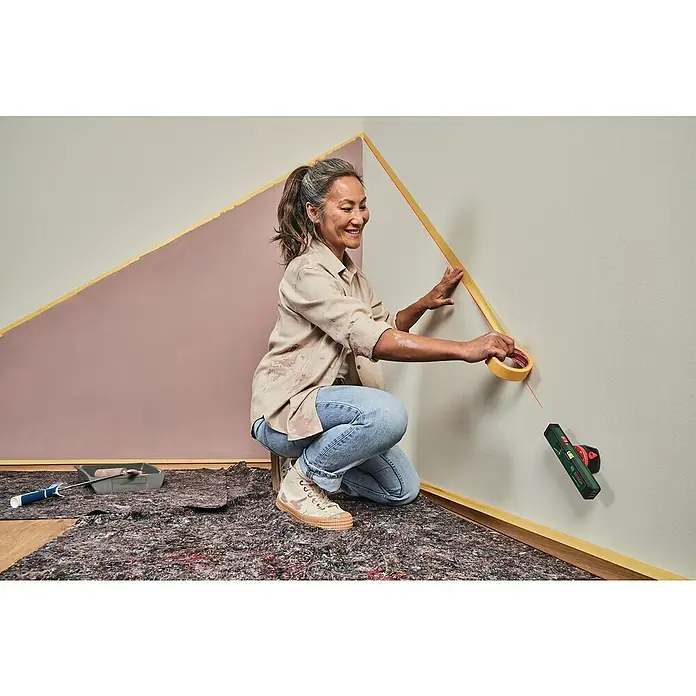Bosch Linienlaser Universal Wall Level - Max. Arbeitsbereich: 5 m - 20 m - Eine Frau bringt mit einem Bosch-Laser Nivelliergerät Malerband an einer Wand an, um ein geometrisches Muster zu erstellen.