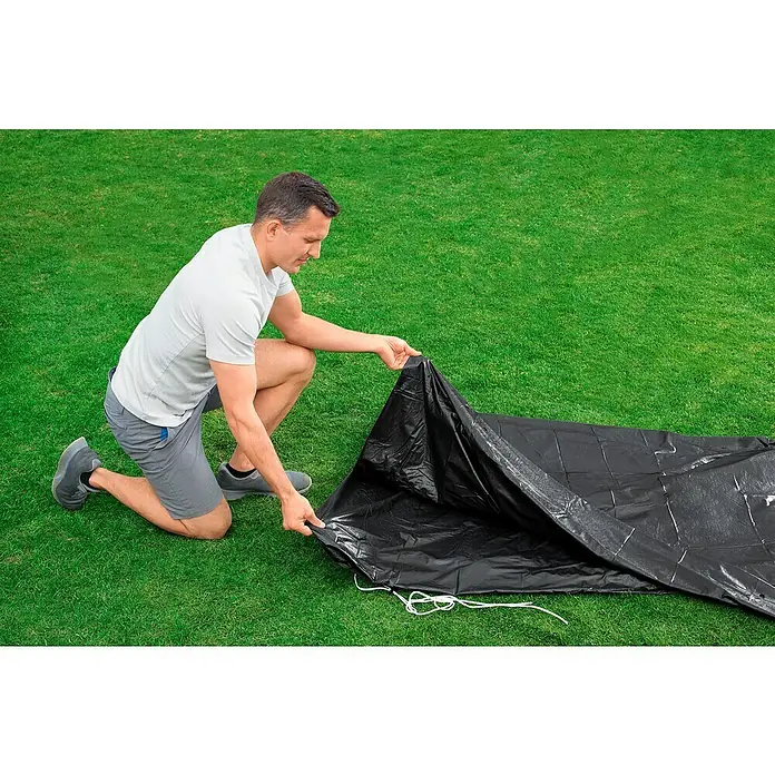 Bestway Pool-Abdeckplane - Kunststoff - Ein Mann kniet auf dem Rasen und breitet eine schwarze Abdeckplane mit weißen Schnüren aus, um den Pool zu schützen.